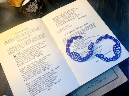 Resting inn open old book of poems are two R+D floral hoop earrings. They are a rich purple color with leaves and blooms twisting around to make a partial wreath shape.