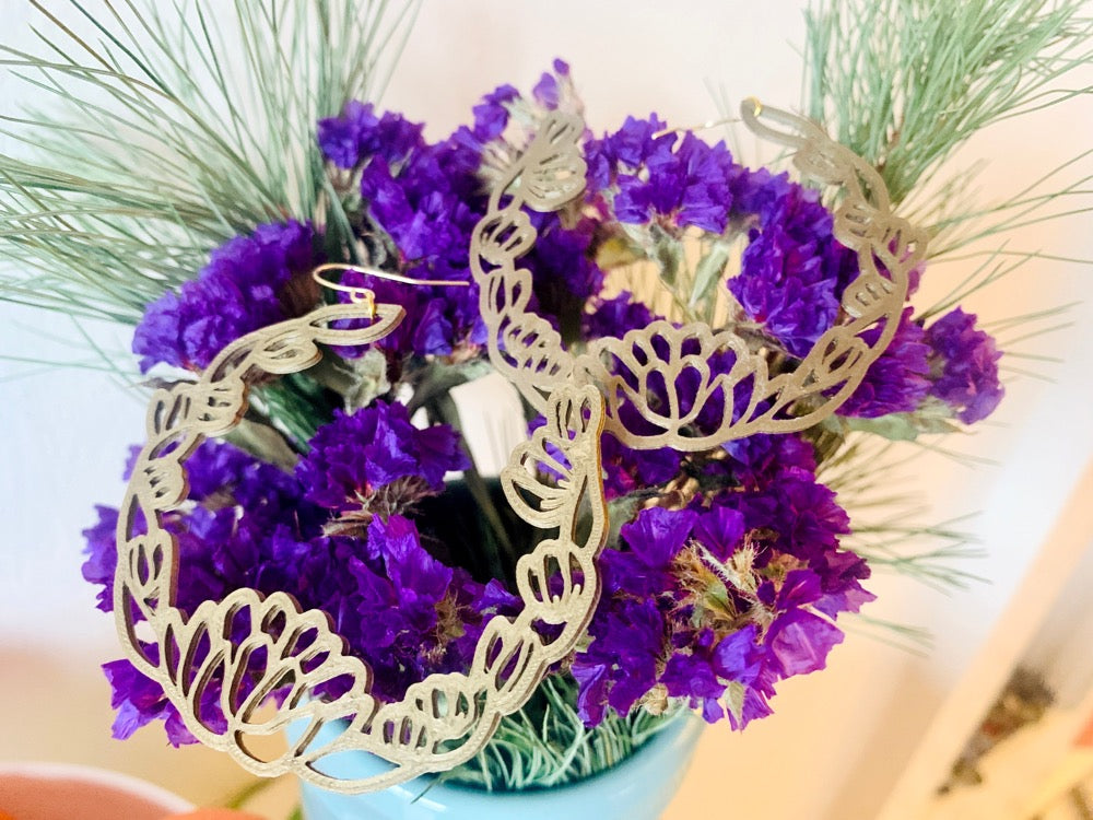 Resting on a bundle of dried purple flowers are two R+D 3D Printed earrings. They are gold in color and shaped as floral wreaths with leaves and blooms layered together to make a circle. 