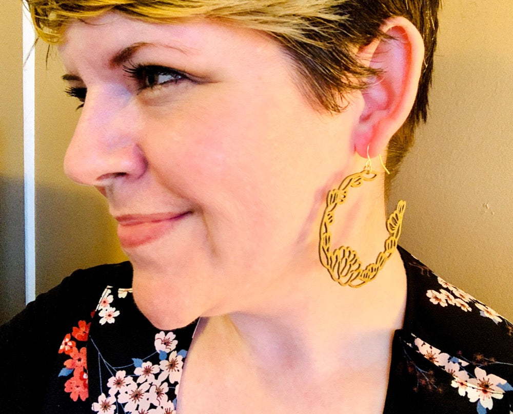 A woman is shown in profile looking away from the camera wearing a R+D 3D printed earring. It is a gold filament with blooms and leaves layered together to form a floral wreath hoop. 