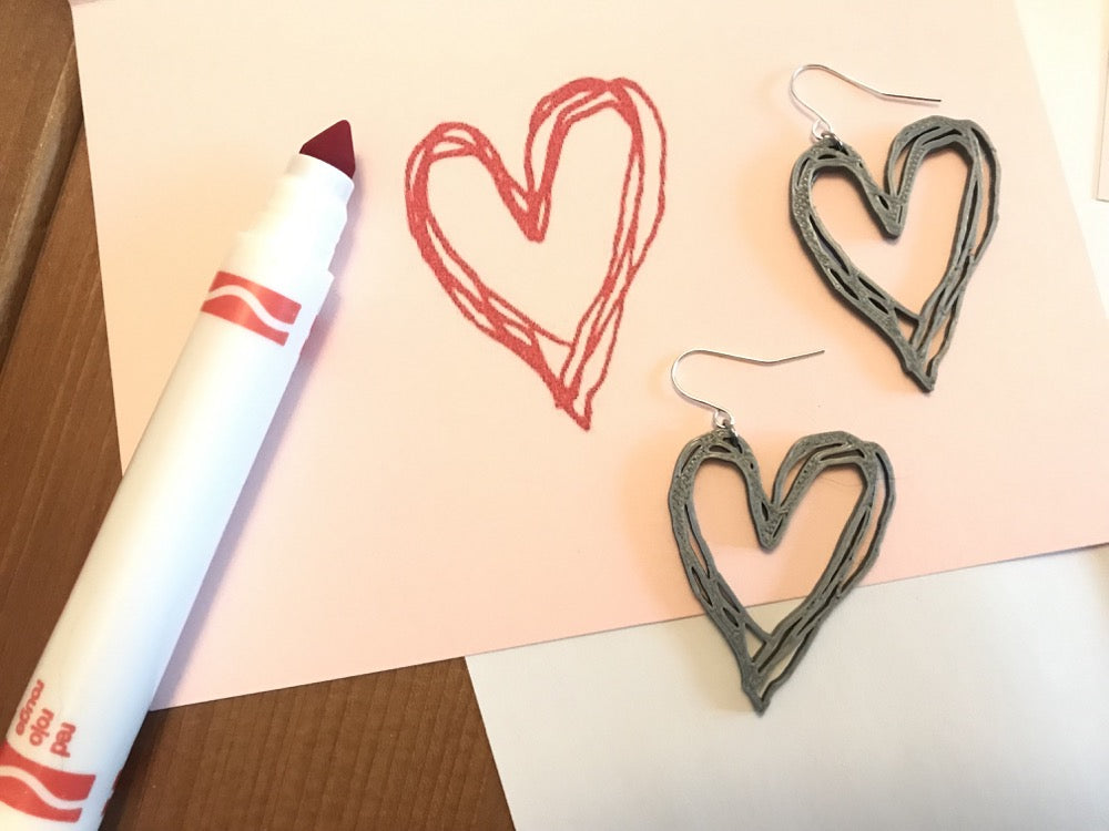 There is a pink card with a uncapped red marker. On the card is a heart drawn with the marker. There are two earrings that are in the same shape as the heart drawing. They are 3D printed with a silver plant based filament.
