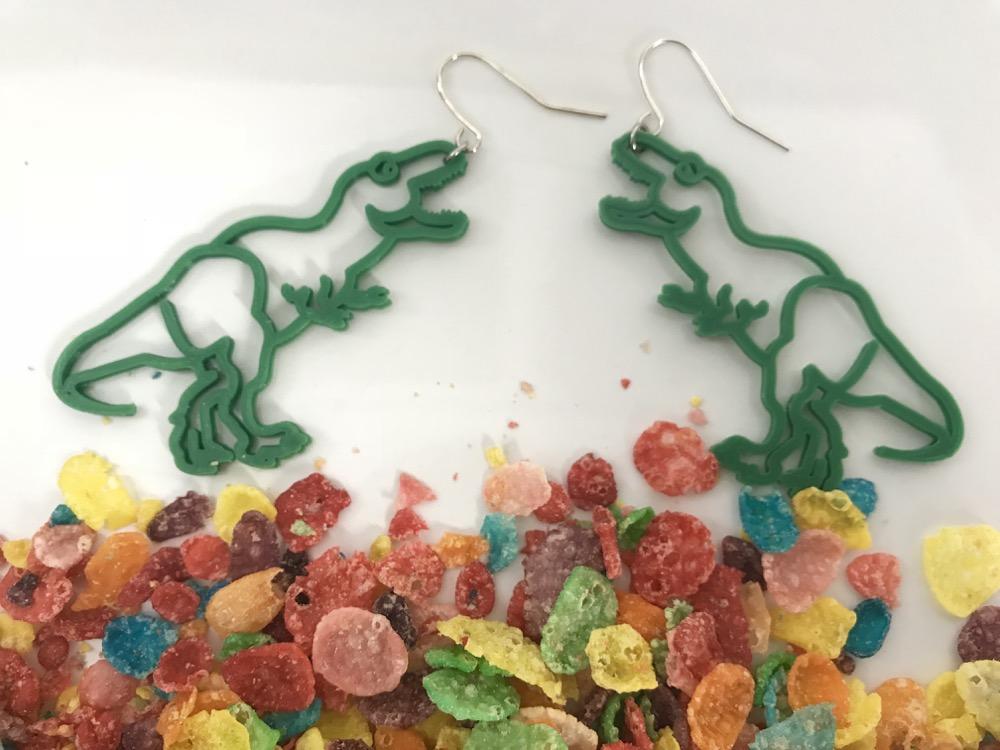 Above what looks like rubble or stones (but is actually fruity pebbles) are two earrings shaped like dinosaurs, specifically like t-rex that are facing each other. The dinosaur earrings are a bright kelly green color and 3D printed in a sustainable plant based filament. 