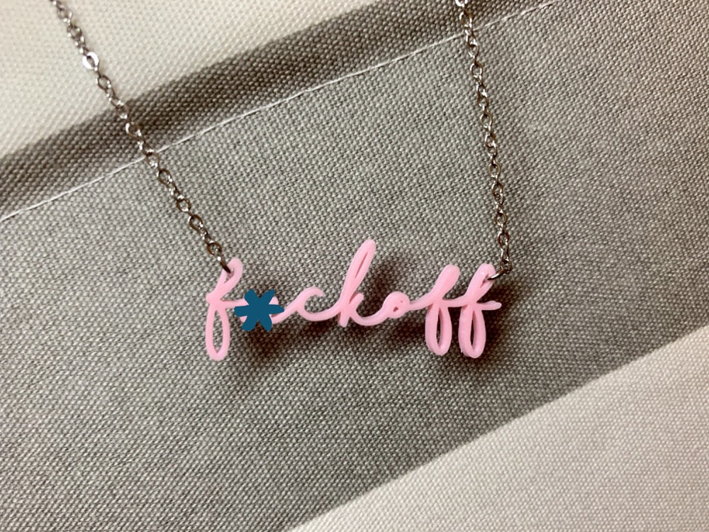 On a grey and white striped background is a  is a 3D printed pendant. In a modern cursive script it says f*ckoff in a light pink color. There is an asterisk over the u in the photo.