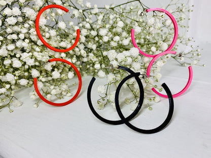 On a white background are springs of baby's breath flowers with three pairs of 3D printed hoop earrings hanging off of them. They are lightweight, plant based, and durable. These are shown in a bright red, black, and hot pink.
