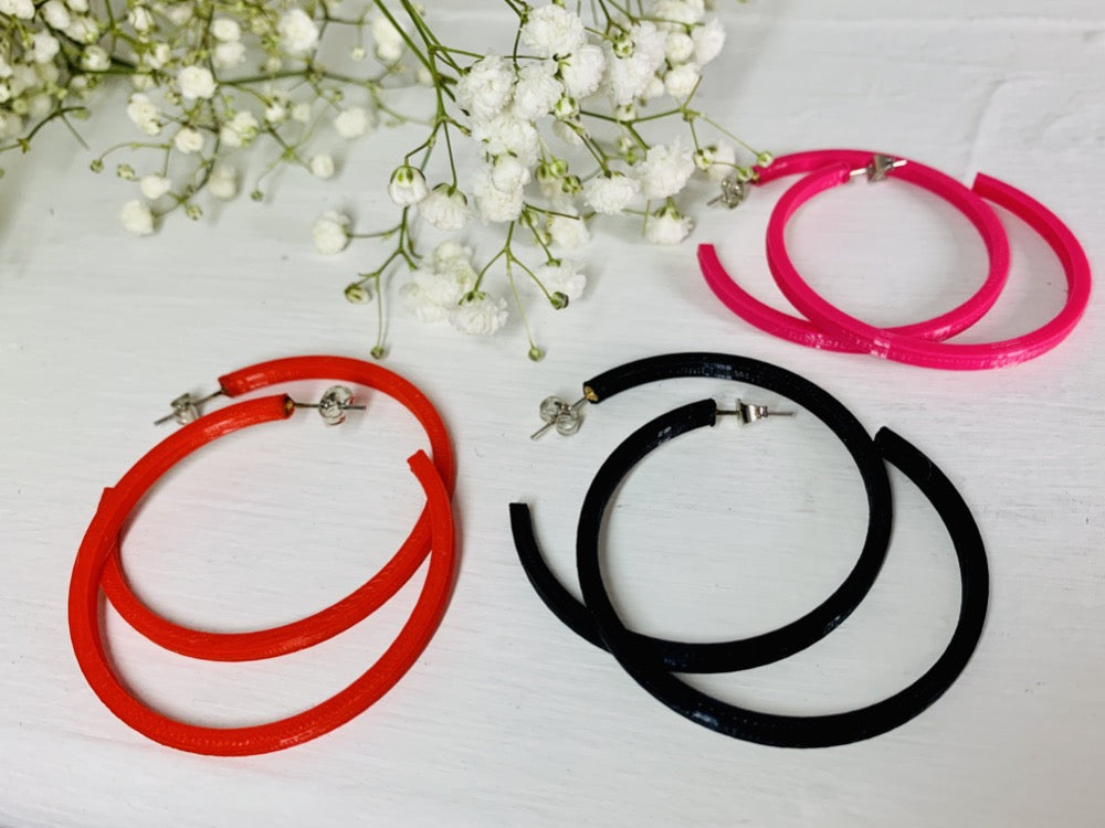 On a white background with baby's breath flowers in the corner are three 3D printed hoop earrings. They are lightweight, plant based, and durable. These are shown in a bright red, black, and hot pink. 