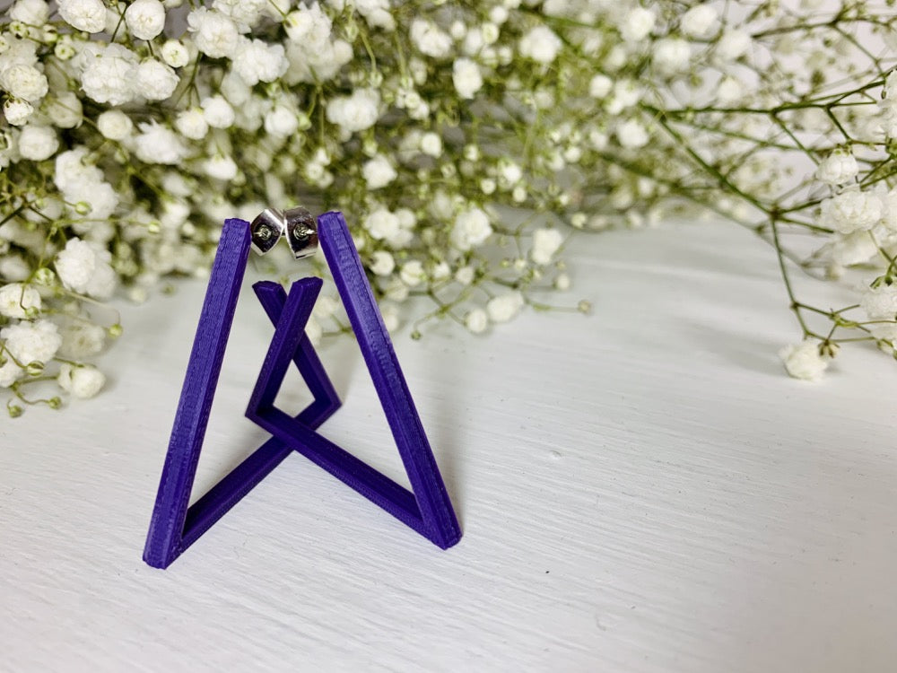 The background is filled with baby's breath flowers. In the foreground there are two 3D printed earrings. They are printed in a plant based purple filament and are shaped as triangle hoops. In the photo they are standing up to create a unique geometric structure. 