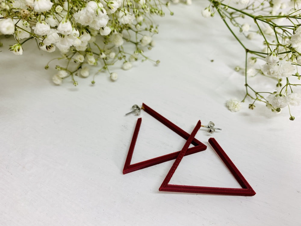 Laying on a white background are two R+D 3D printed earrings. They are triangle hoops that are printed in a plant based filament that is a deep red or merlot color. On the edges of the photo springs of baby's breath flowers are laying down