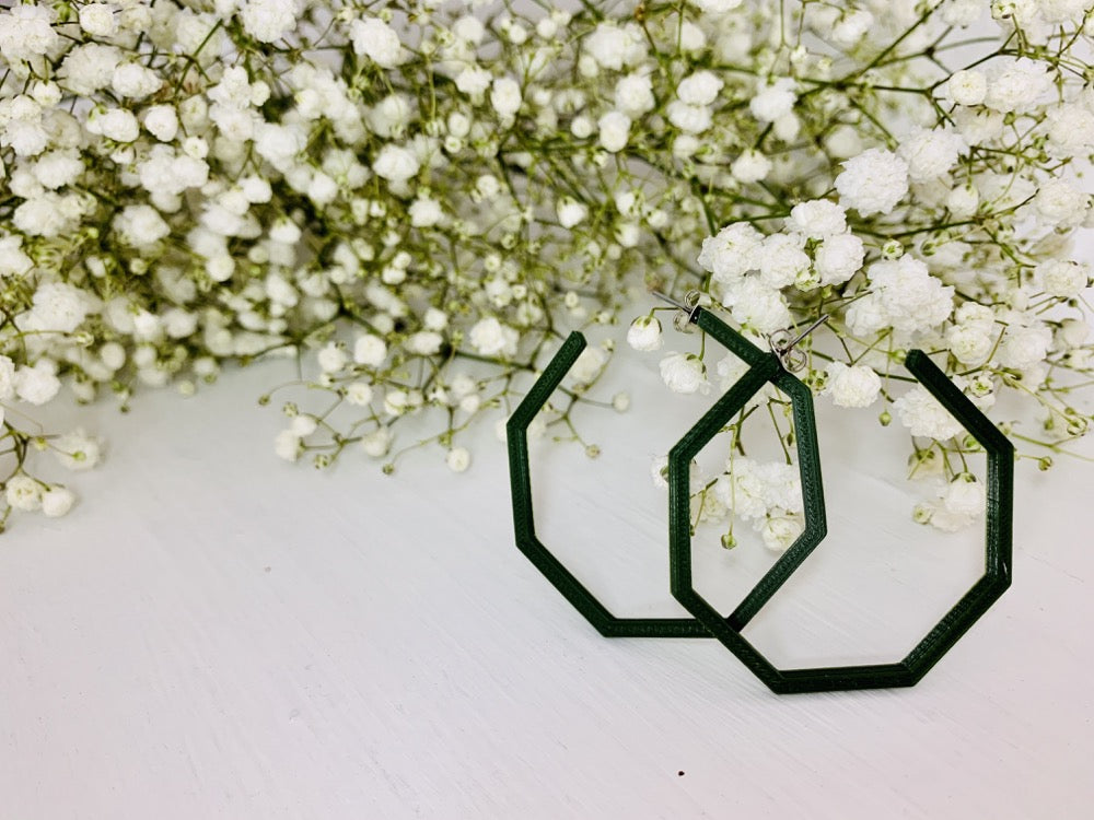 In front of many sprigs of baby's breath are two 3D printed earrings. The earrings are large hoops in the shape of hexagons. 