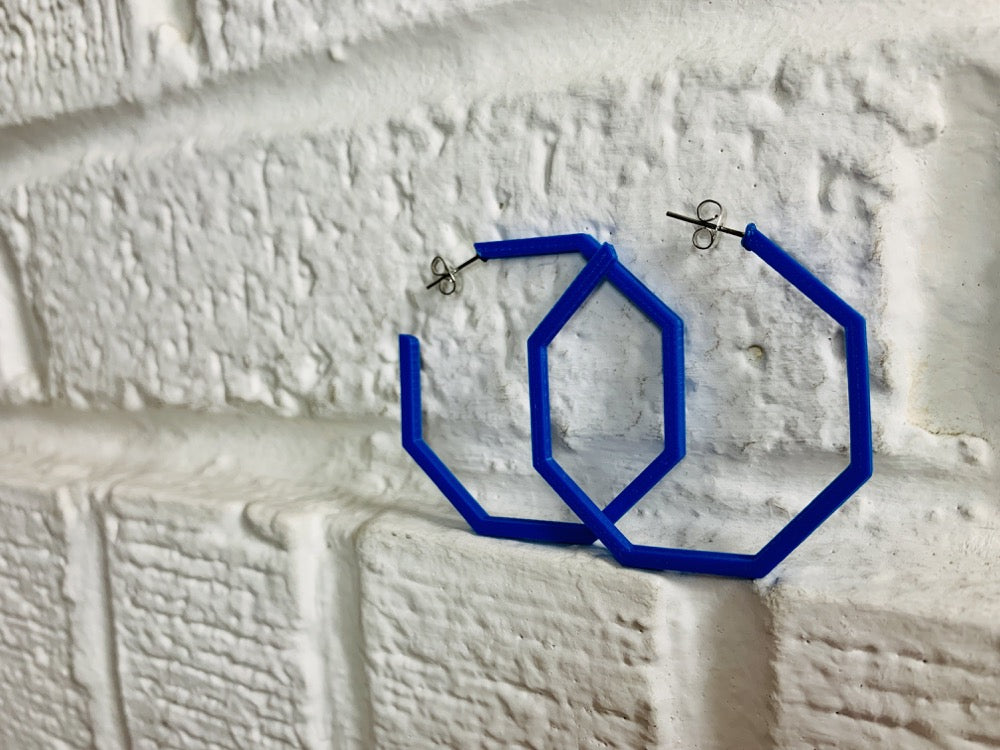 In front of a white brick wall are two 3D printed hoop earrings. They are in the shape of hexagons and are printed in a royal blue with a plant based filament. 