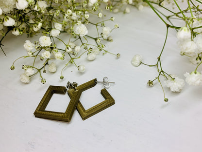 On a white background and surrounded by sprigs of baby's breath flowers are two square hoops that are 3D printed in a gold plant based filament. 