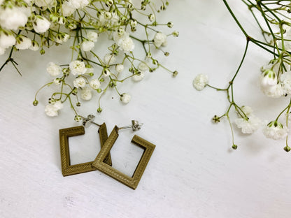 On a white background and surrounded by sprigs of baby's breath flowers are two square hoops that are 3D printed in a gold plant based filament. 