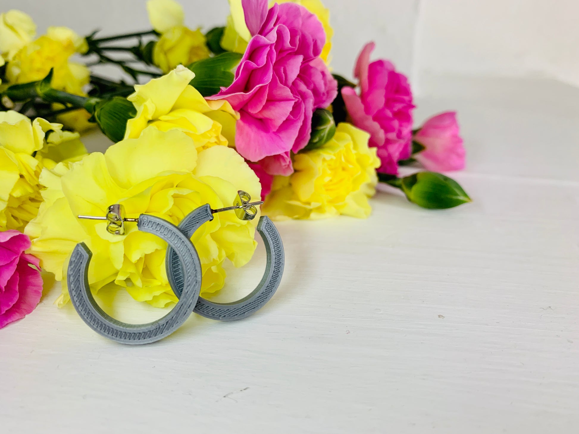 Resting in front of yellow and pink carnations are two 3D printed hoops. They are silver and chunky in their shape. 