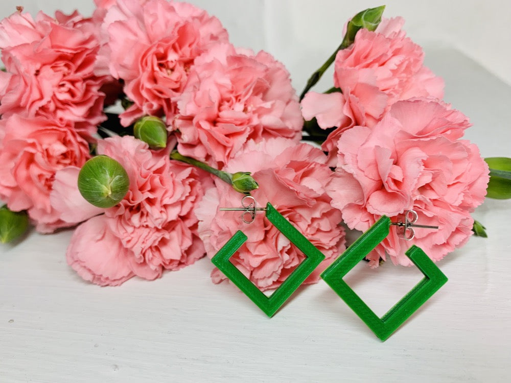 On a white background and surrounded by bright pink carnations are two square hoops that are 3D printed in a kelly green plant based filament. 