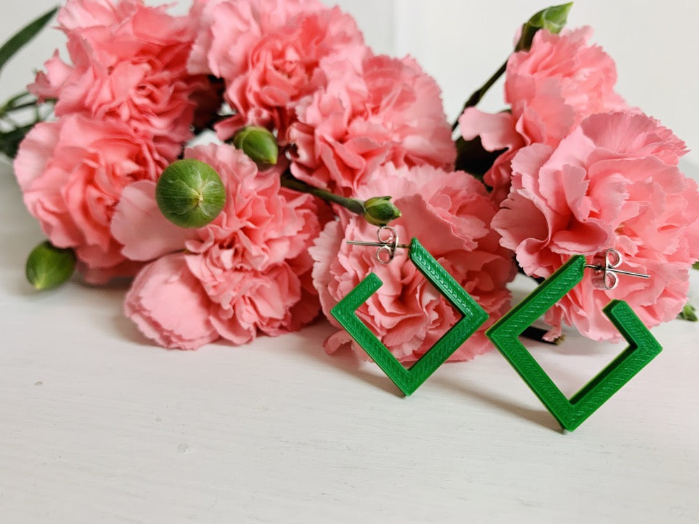 On a white background and surrounded by bright pink carnations are two square hoops that are 3D printed in a kelly green plant based filament.