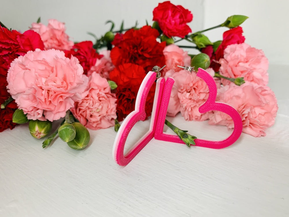 Resting in front of pink and red carnations are two 3D printed hoops from R+D. The hoops are in the shape of hearts with 3 colors layered on top of one another: white, light pink and hot pink. 