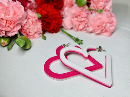Laying on a white background are two 3D printed hoop earrings from R+D. They are layered with three colors: white, light pink, and hot pink. One is flipped over so that the color variations are visible. In the background are bunches of red and pink carnations with green buds reaching out of the cluster.