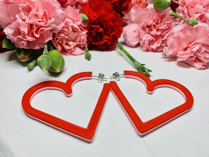 Laying on a white background are two 3D printed hoop earrings from R+D. They are layered with three colors: Light pink, white, and red. In the background are bunches of red and pink carnations with green buds reaching out of the cluster. 