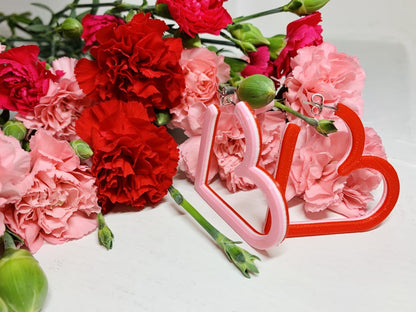 Resting in front of pink and red carnations are two 3D printed hoops from R+D. The hoops are in the shape of hearts with 3 colors layered on top of one another: light pink, white, and red. One of the hoops is turned so that you can see the three colors of the earring. 