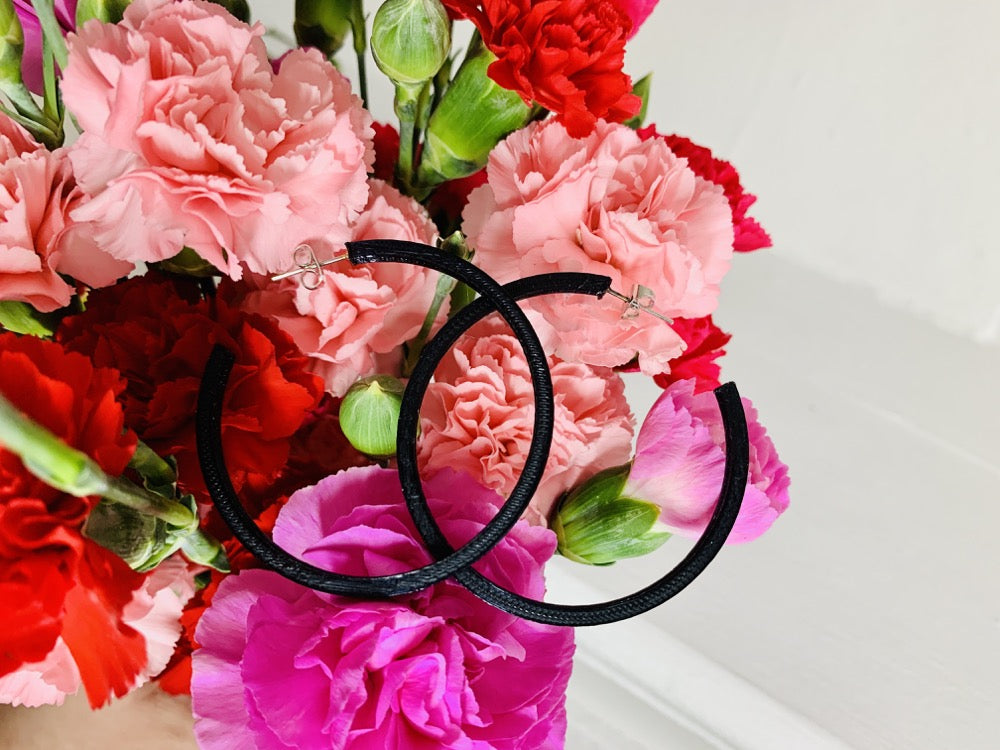 In the background is a bouquet of pink, red, and fuchsia carnations. Resting on the bouquet are two 3D printed hoop earrings. They are black, lightweight, and made from a plant based filament.