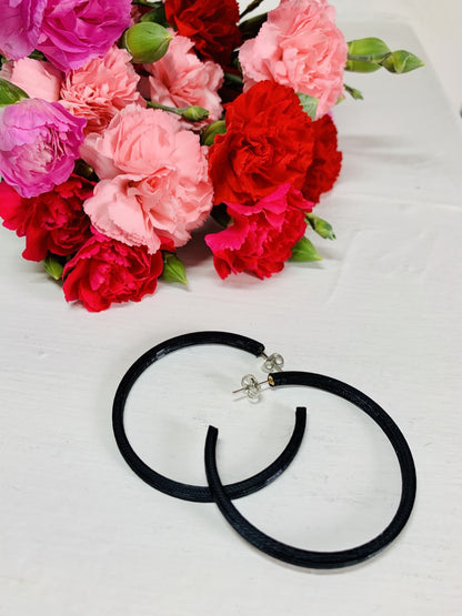 In the background is a bouquet of pink, red, and fuchsia carnations. In the foreground are two 3D printed hoop earrings. They are black, lightweight, and made from a plant based filament. 