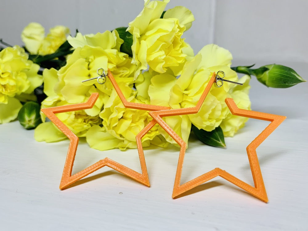 Resting in front of bright yellow carnations are two 3D printed hoop earrings. They are orange and shaped like stars with 5 points. 