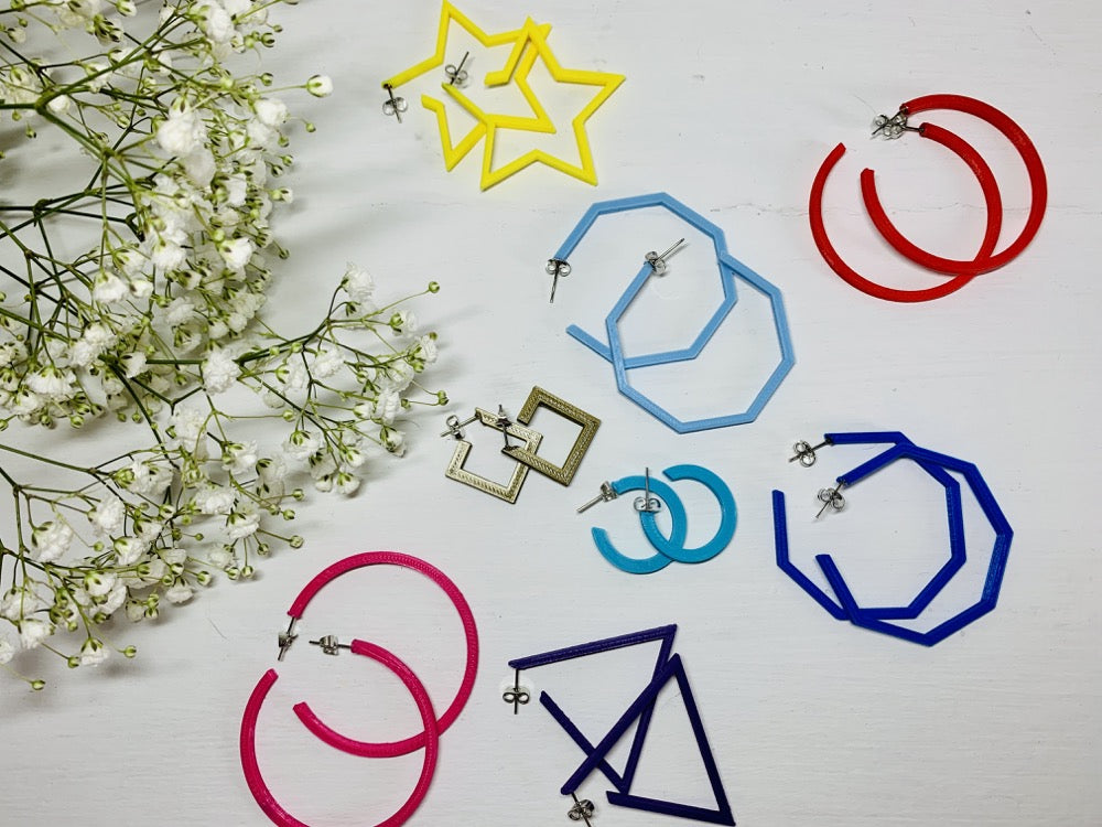 In the upper corner is a sprig of baby's breath. In the foreground there are 8 different pairs of R+D 3D printed hoops. They range in colors from hot pink and red to blues, purple, and gold. They also range in shapes. There are hexagons, smooth hoops, triangles, squares, and stars.