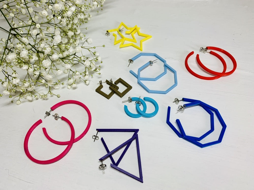 In the upper corner is a sprig of baby's breath. In the foreground there are 8 different pairs of R+D 3D printed hoops. They range in colors from hot pink and red to blues, purple, and gold. They also range in shapes. There are hexagons, smooth hoops, triangles, squares, and stars. 