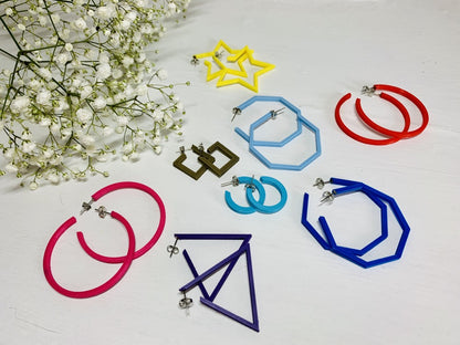 In the upper corner is a sprig of baby's breath. In the foreground there are 8 different pairs of R+D 3D printed hoops. They range in colors from hot pink and red to blues, purple, and gold. They also range in shapes. There are hexagons, smooth hoops, triangles, squares, and stars. 