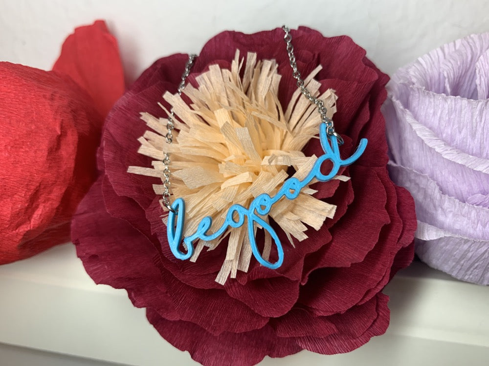 Hanging on a paper flower that is a dark red with a creamy peach center is a 3D printed pendant. In a modern cursive script it says be good  in a bright teal color.