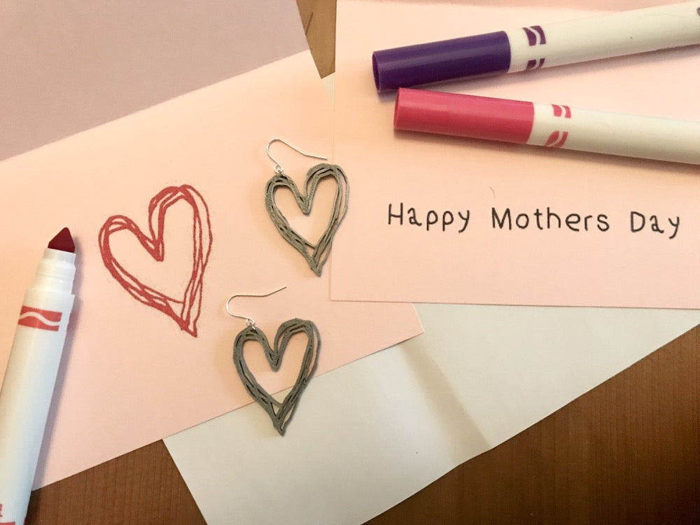 There is a pink card with a uncapped red marker. On the card is a heart drawn with the marker. There are two earrings that are in the same shape as the heart drawing. They are 3D printed with a silver plant based filament.  Another card next to it says Happy Mother's Day.