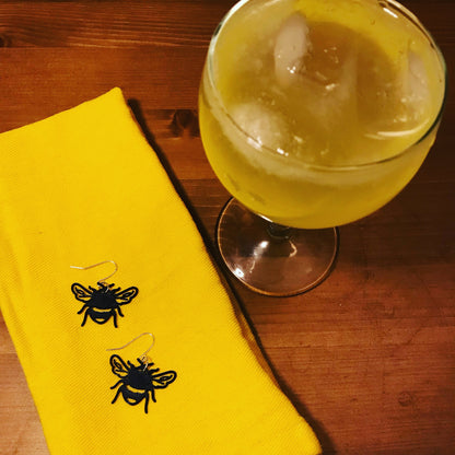 There is a light yellow drink in a coup glass. It is a gin based drink called a bee's knees. Next to it on a wooden counter is a bright yellow cloth napkin. On the napkin are two R+D earrings. They are shaped like bumble bees straight on; they have delicate veins in the wings and the body includes the quintessential stripes of a bee that are fuzzy for collecting pollen.