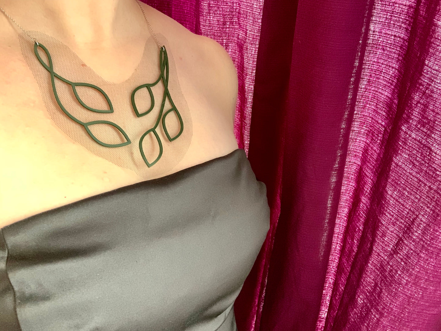 This shows a woman wearing a statement necklace and a satin black strapless dress. The necklace is olive green vines and leaves that twist into an elegant v shape. The piece is 3D Printed with sustainable plant based filament. It is olive green and embedded with a black tulle fabric so that there is a gap between the leaves and it almost has the effect of floating across one's collar bones. 