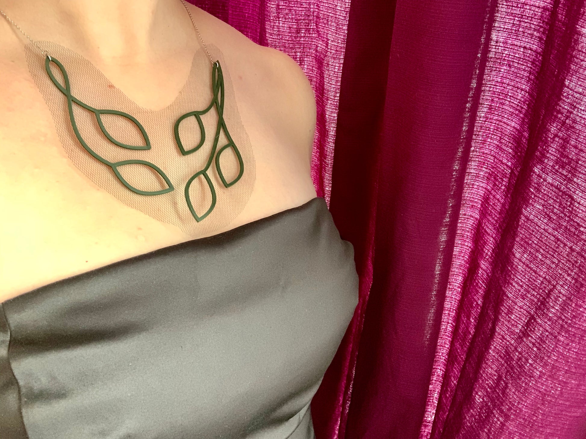 This shows a woman wearing a statement necklace and a satin black strapless dress. The necklace is olive green vines and leaves that twist into an elegant v shape. The piece is 3D Printed with sustainable plant based filament. It is olive green and embedded with a black tulle fabric so that there is a gap between the leaves and it almost has the effect of floating across one's collar bones. 