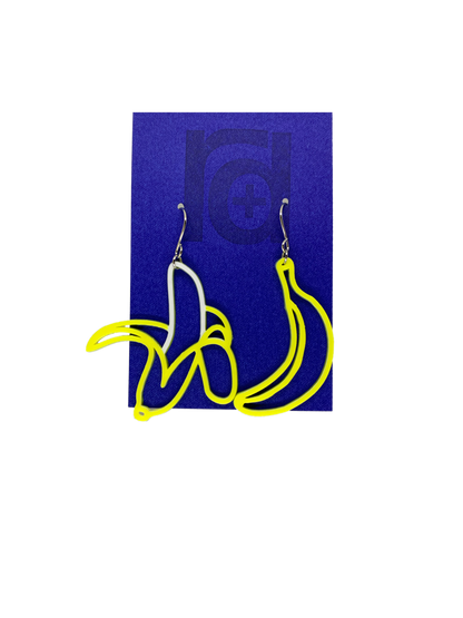 On a bright blue earring card that is the same color as the Chiquita Banana stickers, hang two R+D 3D Printed earrings. They are asymmetrical styles, but both bananas. One is a bright unpeeled, yellow  banana and the other has the skin pulled down to reveal a white banana.