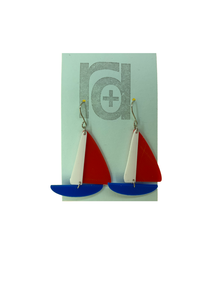 On a light blue earring card are two R+D earrings. They are shaped as sailboats with three pieces: A thin white sail, and larger bright red sail and a classic cobalt blue hull.