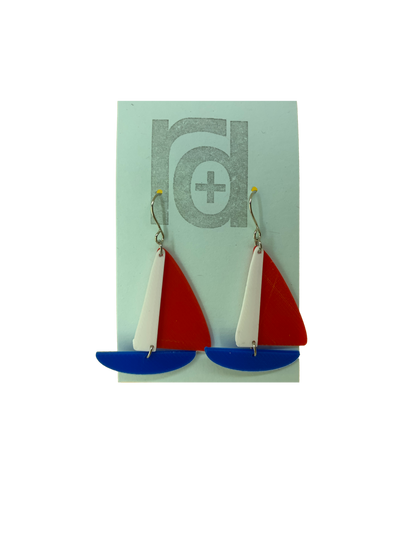 On a light blue earring card are two R+D earrings. They are shaped as sailboats with three pieces: A thin white sail, and larger bright red sail and a classic cobalt blue hull.