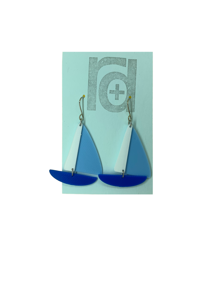 On a light blue earring card are two R+D earrings. They are shaped as sailboats with three pieces: A thin white sail, and larger light blue sail and a classic cobalt blue hull.