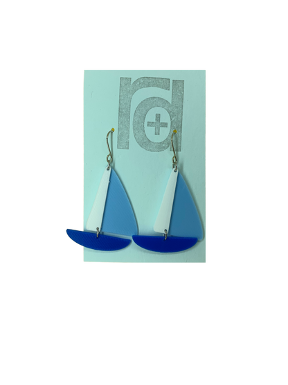 On a light blue earring card are two R+D earrings. They are shaped as sailboats with three pieces: A thin white sail, and larger light blue sail and a classic cobalt blue hull.