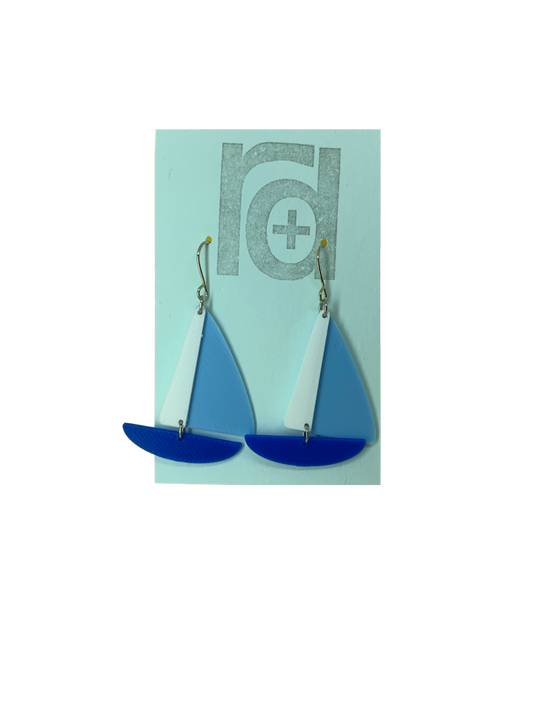 On a light blue earring card are two R+D earrings. They are shaped as sailboats with three pieces: A thin white sail, and larger light blue sail and a classic cobalt blue hull.