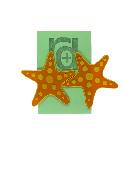 Hanging on a green earring card are two large R+D earrings shaped like star fish. They are orange with a yellow outline and have circle accents across each arm and in the center.