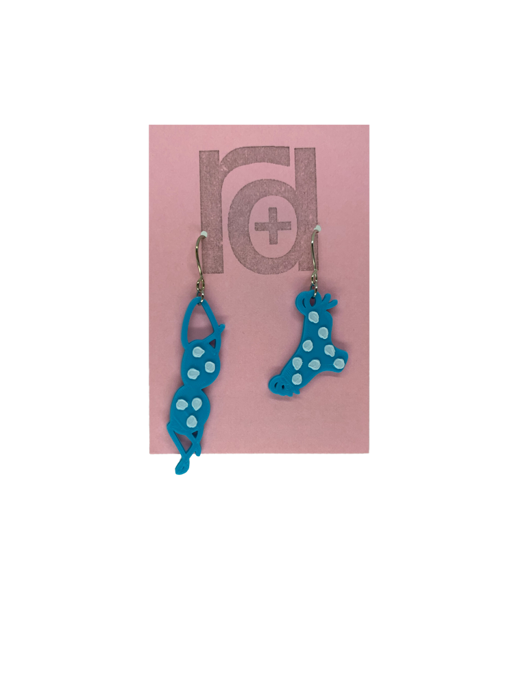 Hanging off of a pink earring card are two R+D earrings. They are asymmetrical  earrings shaped like a classic bikini  that is hung out to dry. The bottoms have ties on the sides. They are teal with white polka dots.
