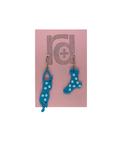 Hanging off of a pink earring card are two R+D earrings. They are asymmetrical  earrings shaped like a classic bikini  that is hung out to dry. The bottoms have ties on the sides. They are teal with white polka dots.