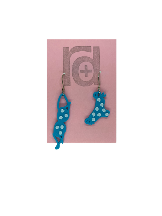 Hanging off of a pink earring card are two R+D earrings. They are asymmetrical  earrings shaped like a classic bikini  that is hung out to dry. The bottoms have ties on the sides. They are teal with white polka dots.