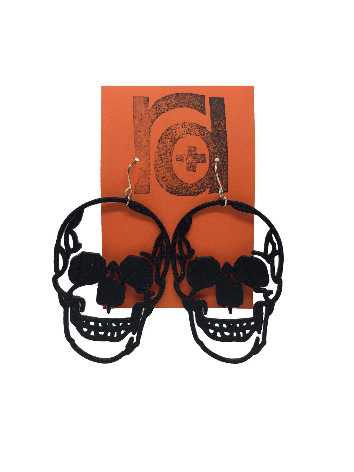 Shown on a orange earring card are two large skull earrings. They are realistic in their shape and are blacked out for the eyes and nose with a jaw that looks like it could be smiling.