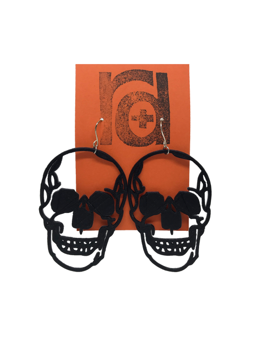 Shown on a orange earring card are two large skull earrings. They are realistic in their shape and are blacked out for the eyes and nose with a jaw that looks like it could be smiling.