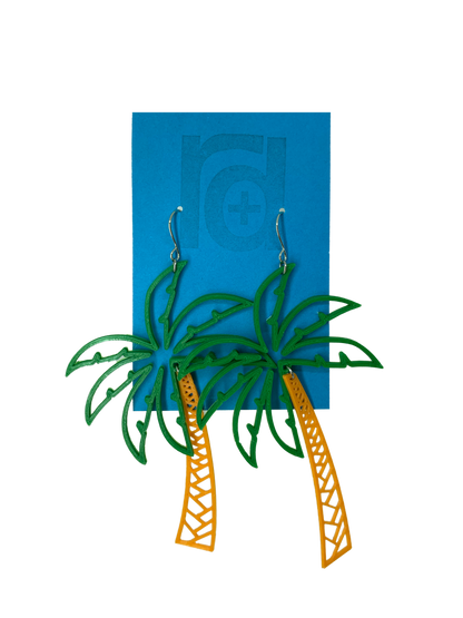 Hanging from a bright blue earring card are two R+D earrings shaped like lush palm trees. They have a wide kelly green fronds above a orange trunk. 
