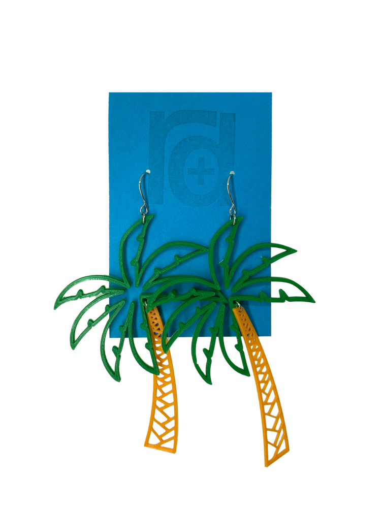 Hanging from a bright blue earring card are two R+D earrings shaped like lush palm trees. They have a wide kelly green fronds above a orange trunk. 