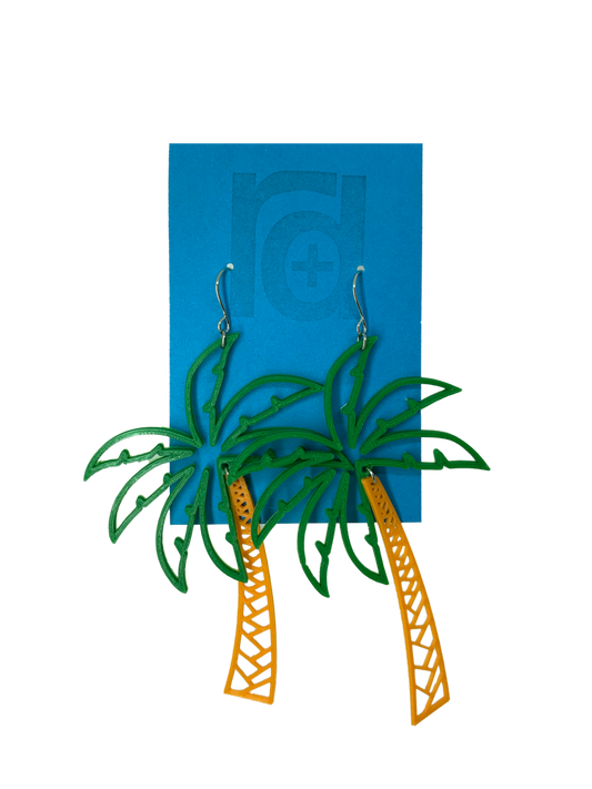 Hanging from a bright blue earring card are two R+D earrings shaped like lush palm trees. They have a wide kelly green fronds above a orange trunk. 