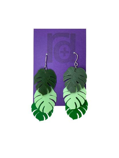 Hanging from a purple earring card are two R+D earrings. The earrings have three monstera frond leaves that hang at different levels. They are also each different color: Olive green, Mint green, and kelly green. 