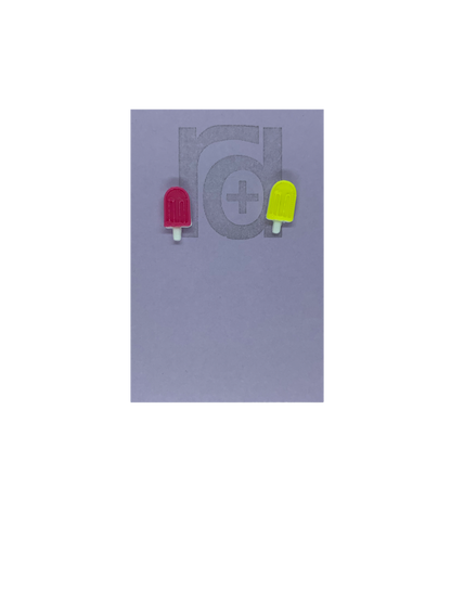 Two small and detailed R+D earrings are shown on a purple earring card. They are shaped as popsicles with white sticks. The earrings are mismatched, one popsicle is hot pink and one is bright yellow.