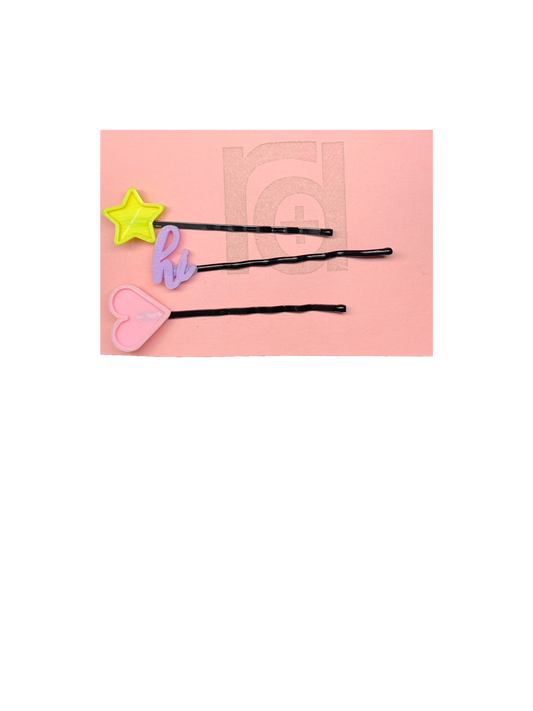 On a pink card are three bobby pins with 3D printed charms on the end from R+D. The  top bobby pin has a bright yellow star. The second is light purple and in cursive script says hi. The third is a light pink heart. All of the packaging is eco friendly, either recyclable or compostable.